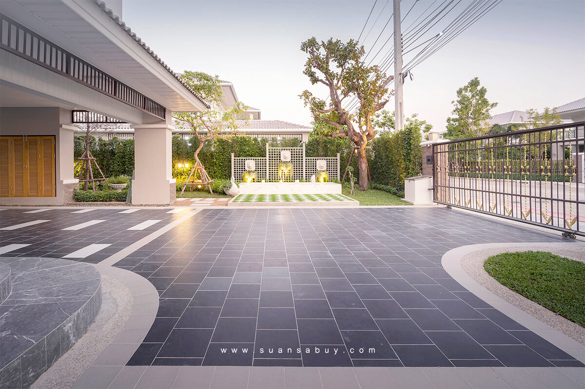 ที่จอดรถปูกระเบื้องผิวด้านสีเข้ม ขอบกรวดล้างสีอ่อน มีวงกลมสนามหญ้าคั่นกลาง มองเห็นทางเดินลายตารางหมากรุกด้านหลัง ผสมผสานวัสดุปูพื้นหลายชนิดให้ที่จอดรถดูสวยงามกลมกลืนกับสวน ผลงานออกแบบโดยสวนสบาย ดีไซน์ สตูดิโอ